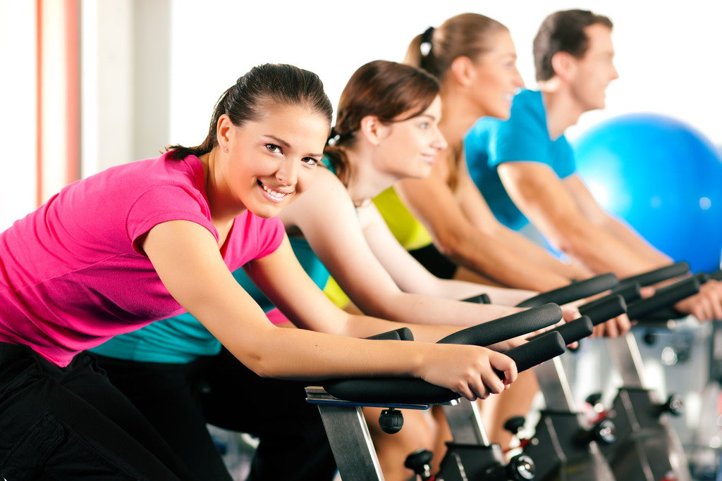 A Woman Smiling While Riding an Exercise Bike With 3 Other People in the Background