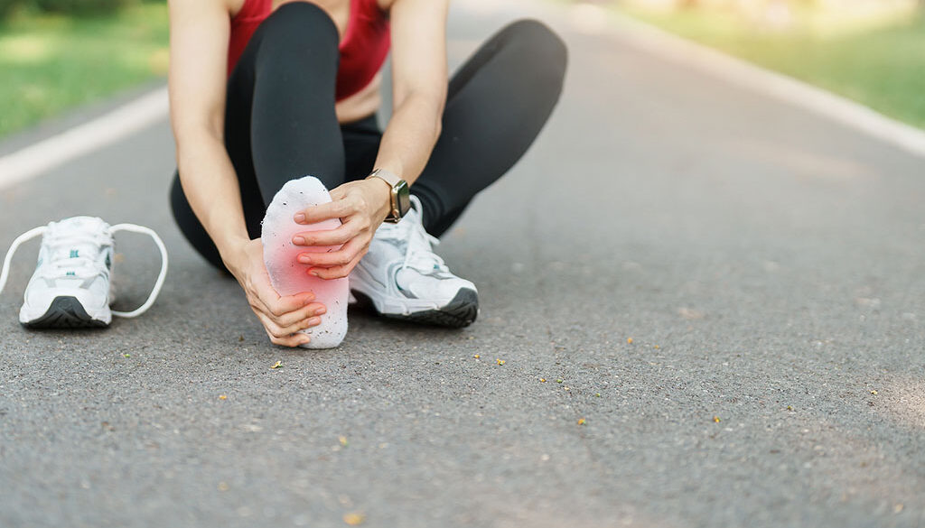 Close Up Of A Runner Sitting On Pavement With Her Shoe Off Grabbing For The Bottom Of Her Foot Experiencing Foot Pain After Running