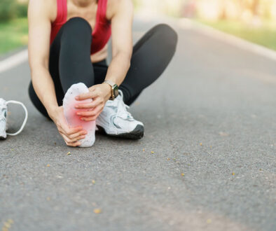 Close Up Of A Runner Sitting On Pavement With Her Shoe Off Grabbing For The Bottom Of Her Foot Experiencing Foot Pain After Running